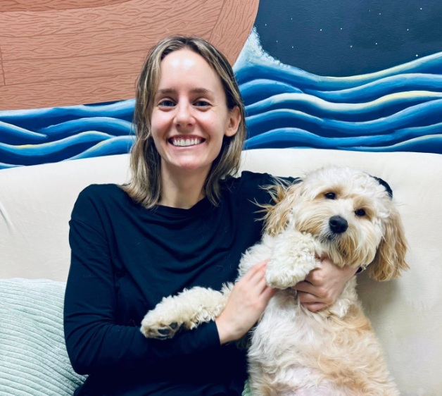 A smiling woman with blonde-highlighted hair wearing a black long-sleeved shirt sits on a white sofa while embracing a fluffy, light-colored cockapoo or goldendoodle dog. The wall behind them features a stylized painted mural of blue ocean waves under a starry night sky.