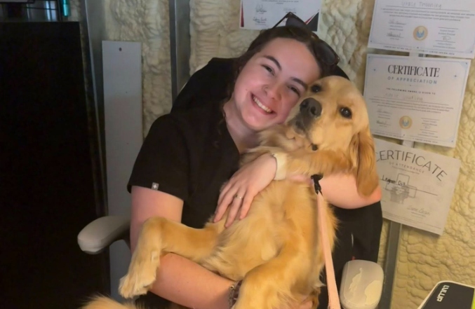 A smiling young woman with dark curly hair wearing black medical scrubs happily cradles a large golden retriever like a baby in an office chair. The friendly dog looks relaxed in her arms, and several framed certificates of appreciation hang on the textured wall behind them.