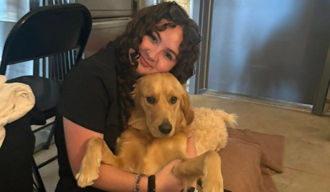 A young woman with curly hair wearing black clothing sits down while holding a golden retriever dog across her lap. She smiles and rests her chin gently on the dog's head, while holding its front paws close. A black folding chair is visible in the background.
