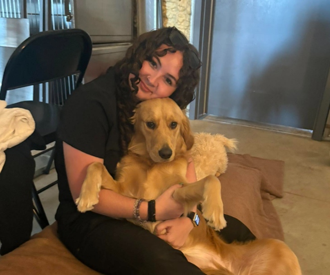 A young woman with curly hair wearing black clothing sits down while holding a golden retriever dog across her lap. She smiles and rests her chin gently on the dog's head, while holding its front paws close. A black folding chair is visible in the background.